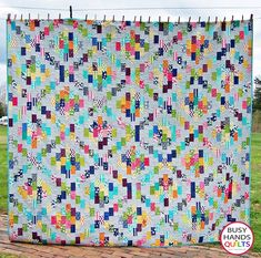 a colorful quilt hanging on the side of a brick wall in front of a grassy field