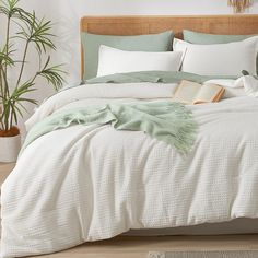 a bed with white sheets and green blankets on it, next to a potted plant