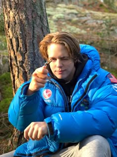 a man in a blue jacket sitting next to a tree and pointing at something with his finger