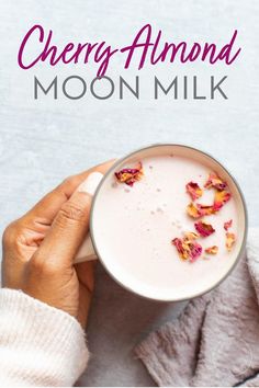 a woman holding a cup of milk with rose petals on it and the words cherry almond moon milk