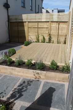 a wooden deck surrounded by plants and flowers next to a building with a fence in the background