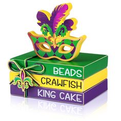 a green box with a purple and yellow mask sitting on top of some books that read beads crawfish king cake