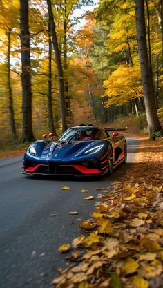 a blue and red sports car driving down a road in the woods with leaves on the ground