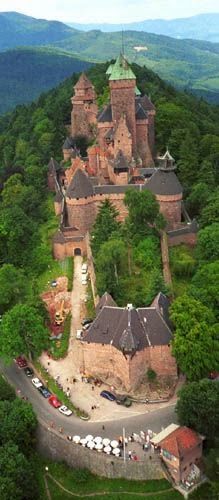 an aerial view of a castle in the middle of trees and mountains with cars parked around it