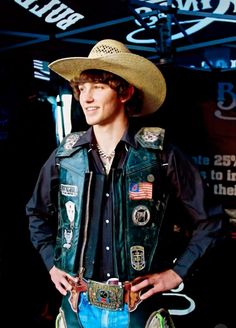 a man wearing a cowboy hat and leather vest with patches on his chest, standing in front of a black background