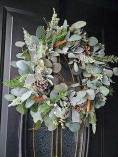 a wreath with greenery and pine cones hanging on a black front door, surrounded by foliage