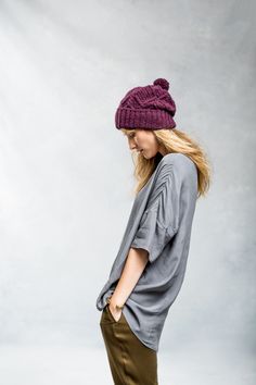 a woman standing in front of a gray wall wearing a purple hat and brown pants