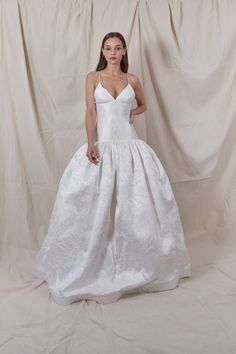 a woman standing in front of a white backdrop wearing a wedding dress with spaghetti straps