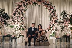 a man and woman sitting on a couch in front of a floral arch with pink flowers