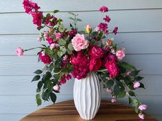 a white vase filled with pink and red flowers on top of a wooden table next to a blue wall