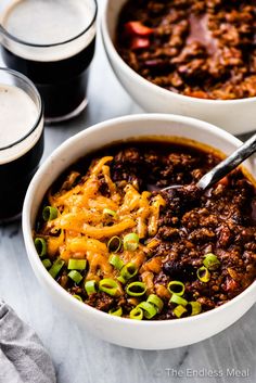 two white bowls filled with chili and cheese next to glasses of cold beer on a table