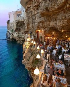 people are sitting at tables on the cliff by the water and eating food in front of them