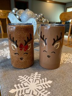 two coffee mugs with reindeer faces on them sitting on top of a table covered in snowflakes