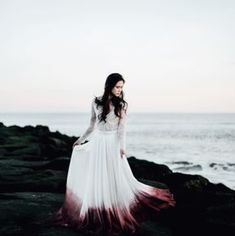 a woman in a white and red dress standing on rocks near the ocean at sunset