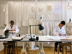 two people working in a sewing studio with lots of white dresses on racks and mannequins behind them