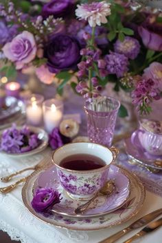 a table set with purple flowers and teacups, candles and plates on it