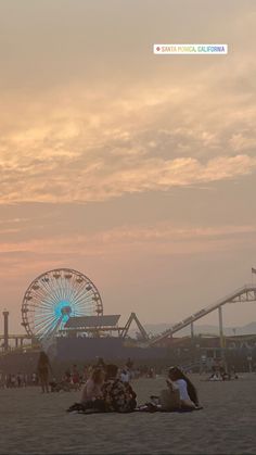 people are sitting on the beach watching the sun go down in front of an amusement park