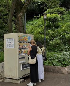 two women standing next to a vending machine in front of a tree and bushes