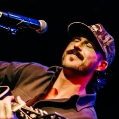 a man with a hat on holding a guitar and singing into a microphone in front of him