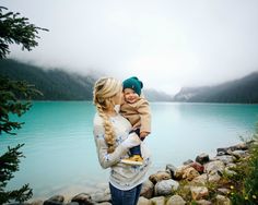 a woman holding a small child in her arms next to a body of water with mountains in the background