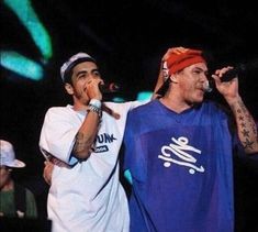 two men standing next to each other on stage with microphones in their hands and one man wearing a red bandana