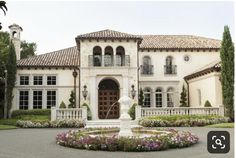 a large house with a fountain in front of it and lots of flowers around the perimeter