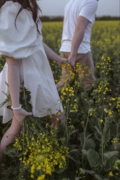 a man and woman walking through a field with yellow flowers in the foreground, holding hands