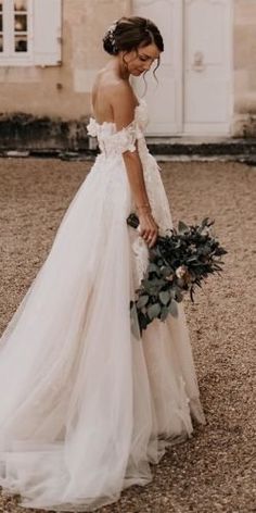 a woman in a wedding dress holding a bouquet and looking down at the ground with an old building behind her