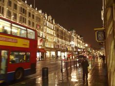 a red double decker bus driving down a street next to tall buildings in the rain