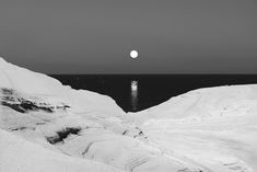 the moon is setting over the ocean in black and white, with snow on the ground