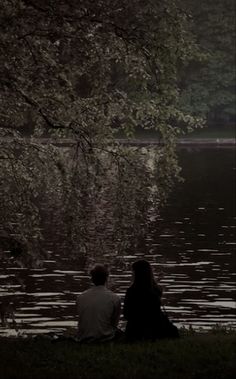 two people sitting on the grass near water under a tree with their backs to each other