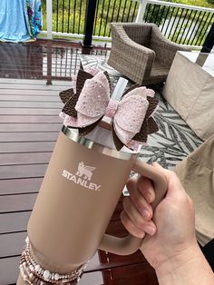 a person holding a coffee mug with pink bows on the top and brown handles, in front of a deck