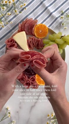 two hands holding slices of ham, cheese and fruit on a plate with flowers in the background