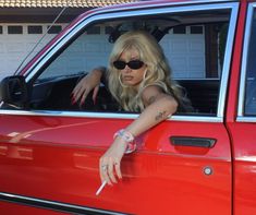 a woman in sunglasses leaning out the window of a red car with her hand on the door handle