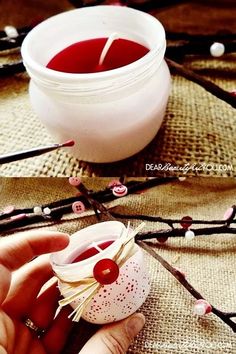 a hand holding a small candle in front of a jar filled with red liquid and cherry blossoms