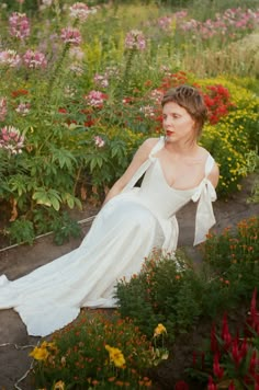 a woman in a white dress is sitting on the ground surrounded by flowers and plants