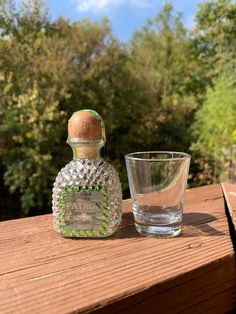 a glass and bottle sitting on top of a wooden table next to each other with trees in the background