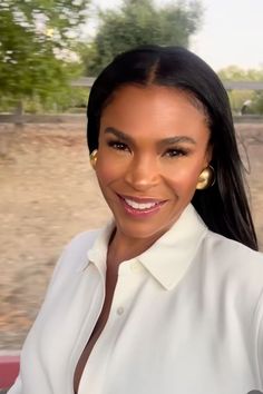 a woman with long black hair and gold earrings smiling at the camera, wearing a white shirt