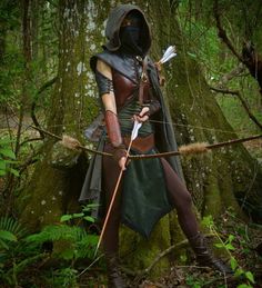 a woman dressed in costume holding an arrow while standing next to a large tree trunk