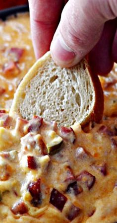 a hand dipping a piece of bread into a casserole with bacon and cheese