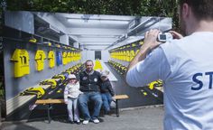 a man taking a photo of two people sitting on a bench in front of a wall with yellow shirts