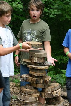 three boys standing around a stack of wooden logs with one boy pointing at the top