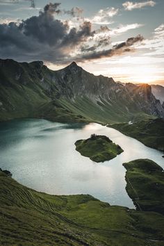 a lake surrounded by green mountains under a cloudy sky with the sun peeking through clouds