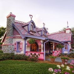 a pink and blue house with lots of flowers on the front lawn, surrounded by greenery