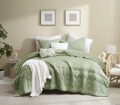 a bed with green comforter and pillows in a room next to a chair, table and potted plant