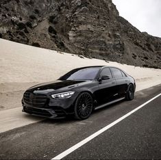 a black car is driving down the road in front of some sand dunes and mountains