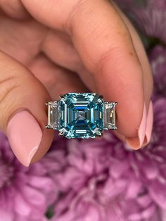 a woman's hand holding an emerald and diamond ring with pink flowers in the background