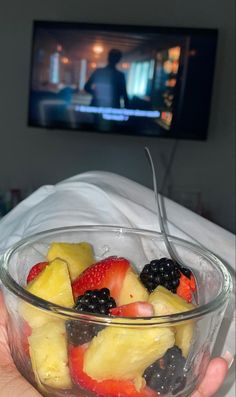 a person holding a bowl of fruit with a television in the backgrouund