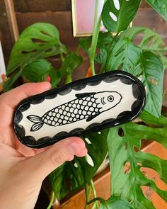 a hand holding up a black and white fish shaped glass vase next to a green plant