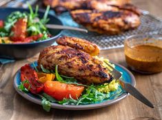 two plates with chicken, vegetables and sauces on them sitting on a wooden table